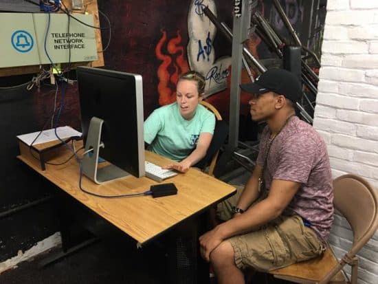 A female volunteer helps a boy in a baseball cap work at a computer on his resume