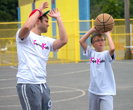 A teenage mentor teaching a young boy how to shoot a basketball during summer camp
