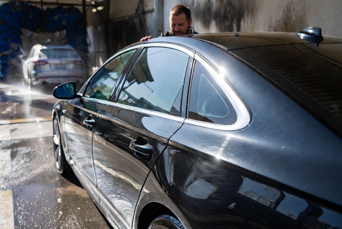automatic brush car wash on a sunny afternoon. In the foreground, a black midsize luxury sedan has just exited the wash and is parked beside the exit lane on wet pavement. The driver, a casually dressed man in his late 30s or early 40s, leans toward the driver's side door, carefully examining the paint with a disappointed expression.