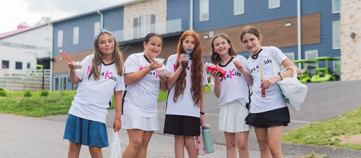 Five girls wearing matching white Kars4Kids T-shirts smile for the camera, holding ice cream, drinks, and snacks in front of a camp building.